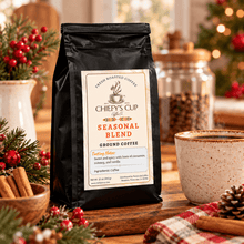 Load image into Gallery viewer, A black bag of Chiefy's Cup Seasonal Blend Ground Coffee stands on a wooden table in a cozy holiday setting, surrounded by a steaming mug, cinnamon sticks, and a blurred Christmas tree in the background.
