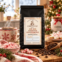 Load image into Gallery viewer, A black bag of Chiefy's Cup Coffee Fresh Roasted Coffee, Frosted Peppermint flavor, stands on a wooden table surrounded by holiday-themed coffee props, candy canes, and a decorated Christmas tree in the background.
