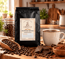 Load image into Gallery viewer, A black bag of Chiefy's Cup Midnight Mocha Ground Coffee stands on a wooden counter in a warm kitchen setting, surrounded by coffee beans, chocolate pieces, a cup of coffee with steam, and bowls of cocoa powder.
