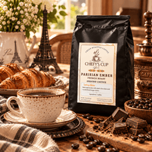 Load image into Gallery viewer, A black bag of Chiefy's Cup Coffee Parisian Ember French Roast stands on a wooden table beside a cup of coffee, chocolate pieces, coffee beans, and croissants, with a Paris-themed decorative scene in the background.
