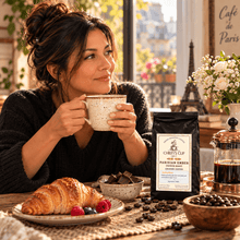 Load image into Gallery viewer, A woman in a cozy kitchen setting holds a speckled mug while a bag of Chiefy's Cup Parisian Ember Medium Roast Arabica Coffee sits on the table next to a croissant, chocolate, and coffee beans.
