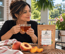 Load image into Gallery viewer, A woman sits on a porch holding a glass of amber tea next to a stand-up pouch of Chiefy's Cup Peach Paradise Green Tea. Fresh peaches and sliced peaches are arranged on a woven table runner in front of her, with greenery and flowers in the background.
