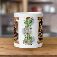 Load image into Gallery viewer, Back side view 11oz white glossy mug with image design of a rustic cowboy hat resting on a cactus, surrounded by small flowering cactus, rocks, desert animals, a cow skull, a wagon wheel, with sparkles and mist in the background.
