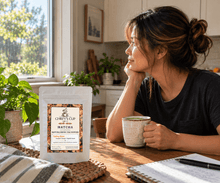 Load image into Gallery viewer, A woman in a kitchen setting looks thoughtfully to the side while holding a mug, with a stand-up pouch of Chiefy's Cup Matcha tea placed on the table in the foreground.
