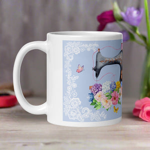 Left side view white glossy mug featuring a vintage sewing machine with a bouquet of flowers, butterflies, and a light blue background framed with lace details.