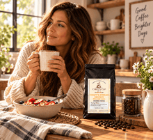 Load image into Gallery viewer, A woman in a cozy knit sweater sits at a wooden table holding a mug, with a bag of Chiefy's Cup Ground Coffee in the foreground and a bowl of fruit and coffee beans on the table. The setting is a warm, sunlit kitchen with shelves and framed wall art.
