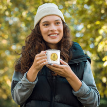Load image into Gallery viewer, A smiling woman outdoors in a forest setting, wearing a cream ribbed beanie, a black puffer vest over a gray hoodie, and holding a white enamel camping mug with a printed map.
