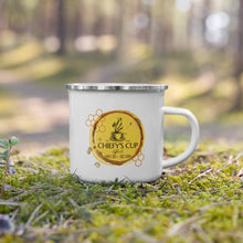 Load image into Gallery viewer, A white enamel camping mug with a silver rim sits on mossy ground in an outdoor forest setting. The mug features a circular yellow and brown graphic with a coffee cup and the text 'CHIEFY'S CUP Cafe'.
