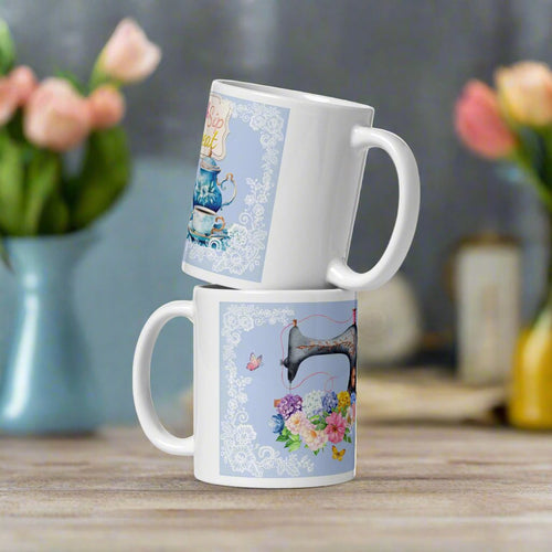 White glossy mug featuring a vintage coffee pot with a cup and saucer on the right, and a vintage sewing machine adorned with a bouquet of flowers and butterflies on the left. Both designs are set against a light blue background framed with delicate lace details, showcasing a charming and elegant theme.