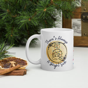 Left side view of a white glossy mug with the phrase "There's Always a Bright Side" in festive font, featuring a cute kitten tangled in Christmas lights, surrounded by a snowflake circle frame and a round yellow hue background with small colorful sparkles.