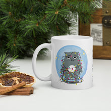 Load image into Gallery viewer, Left side view of a white glossy mug with an image design of a sad kitten tangled in Christmas lights, surrounded by golden sparkles, with a soft blue hue circle background.
