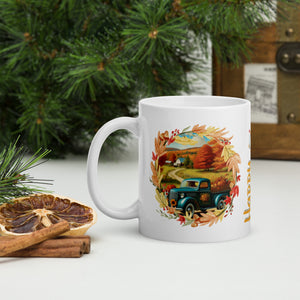 Left side view of a white glossy mug featuring a design of an old blue truck with a bed full of pumpkins on a country road, framed by a vibrant autumn leaf border, set against a late autumn day scenery.