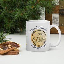 Load image into Gallery viewer, Right side view of a white glossy mug with the phrase &quot;There&#39;s Always a Bright Side&quot; in festive font, featuring a cute kitten tangled in Christmas lights, surrounded by a snowflake circle frame and a round yellow hue background with small colorful sparkles.
